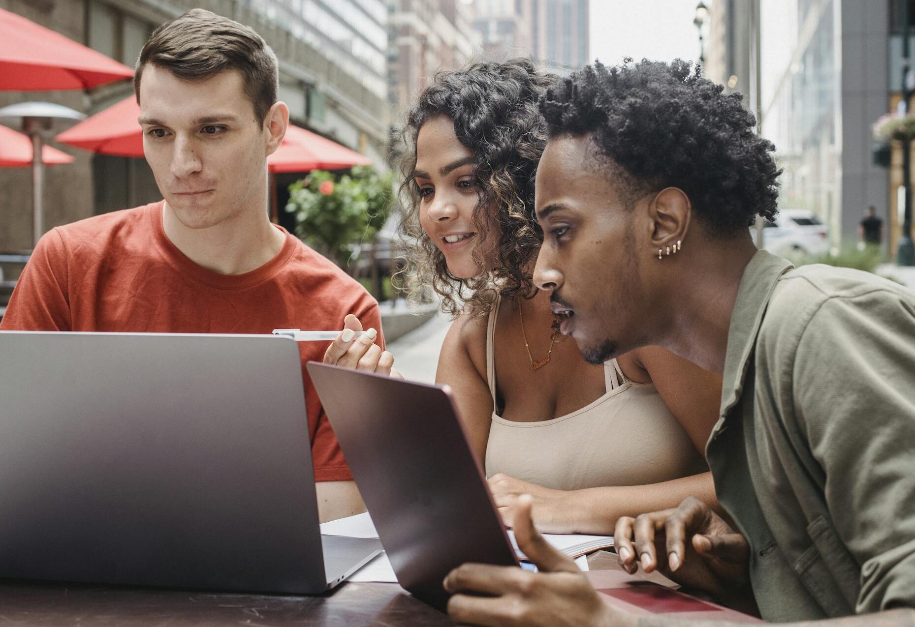 Concentrated young multiracial students sitting in street cafe and using laptops while preparing for exams on sunny day